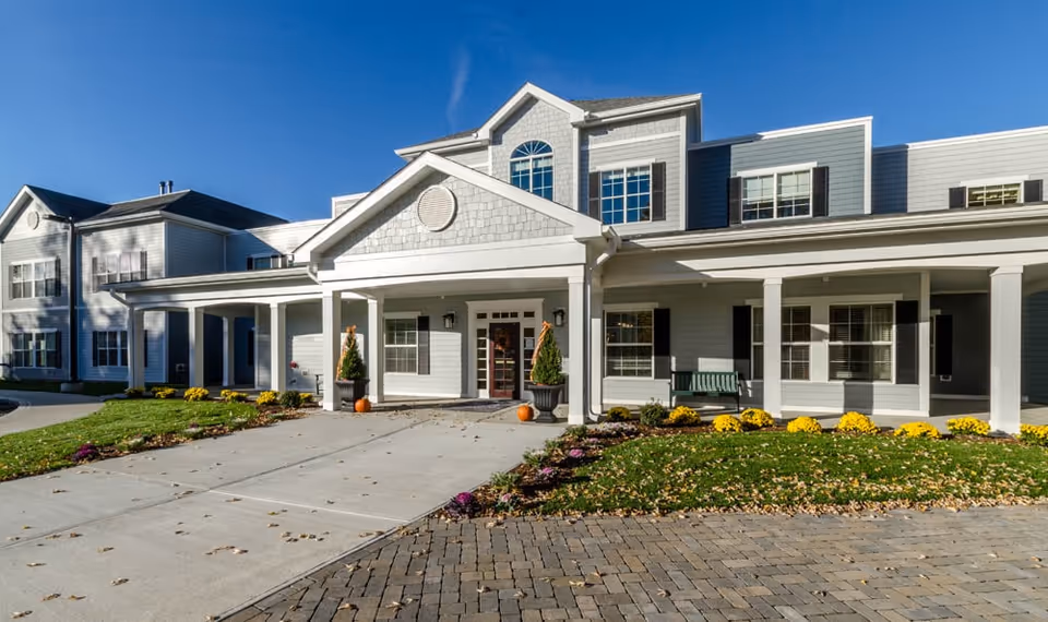 Front exterior view of a senior living facility building with a covered entrance, large windows, and a paved driveway. The building is light gray with white trim and has landscaping with yellow flowers and pumpkins near the entrance.