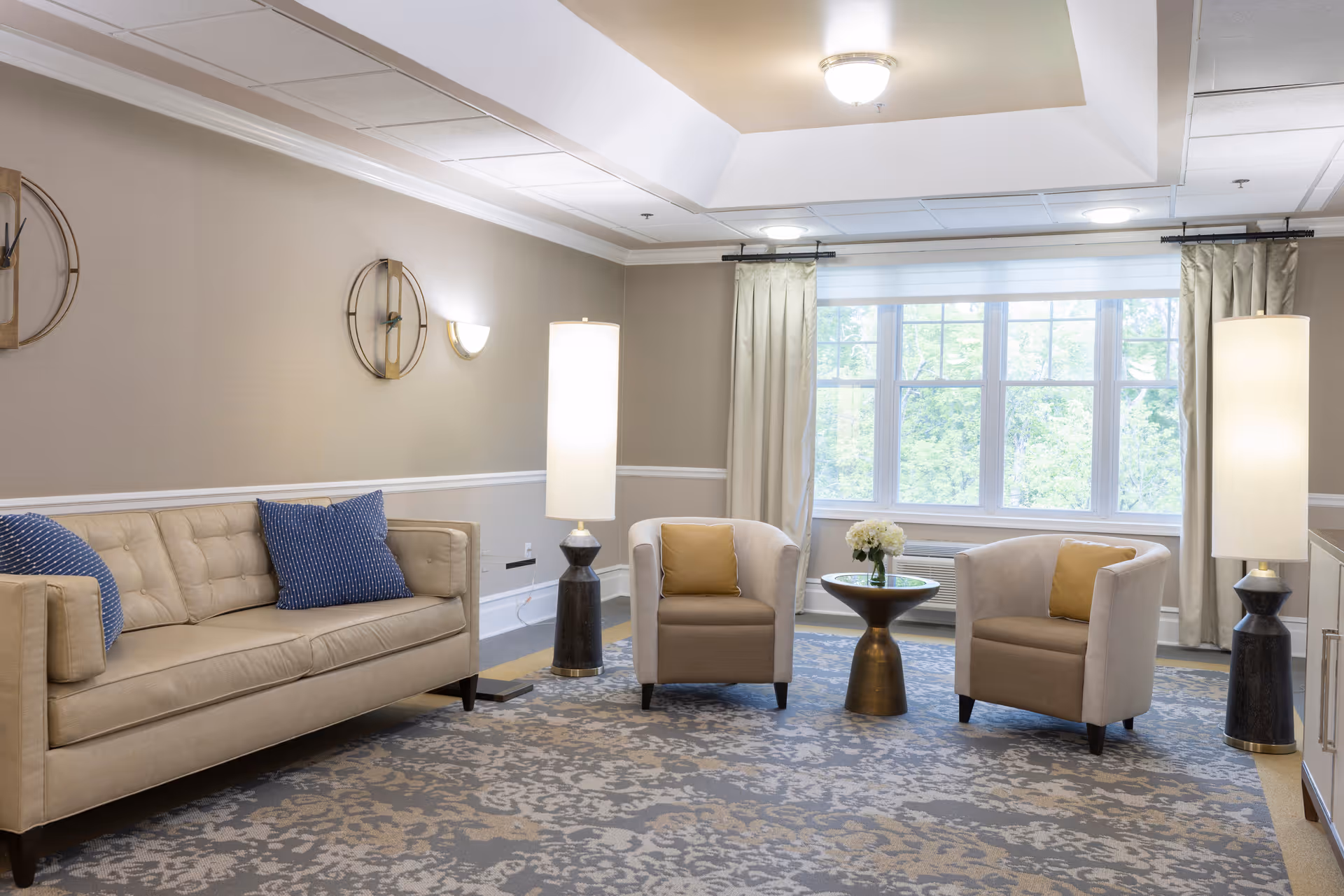 A bright and cozy living room with a beige sofa adorned with blue pillows, two beige armchairs with yellow cushions, a small round table with a flower arrangement, two tall floor lamps, and large windows with curtains letting in natural light.