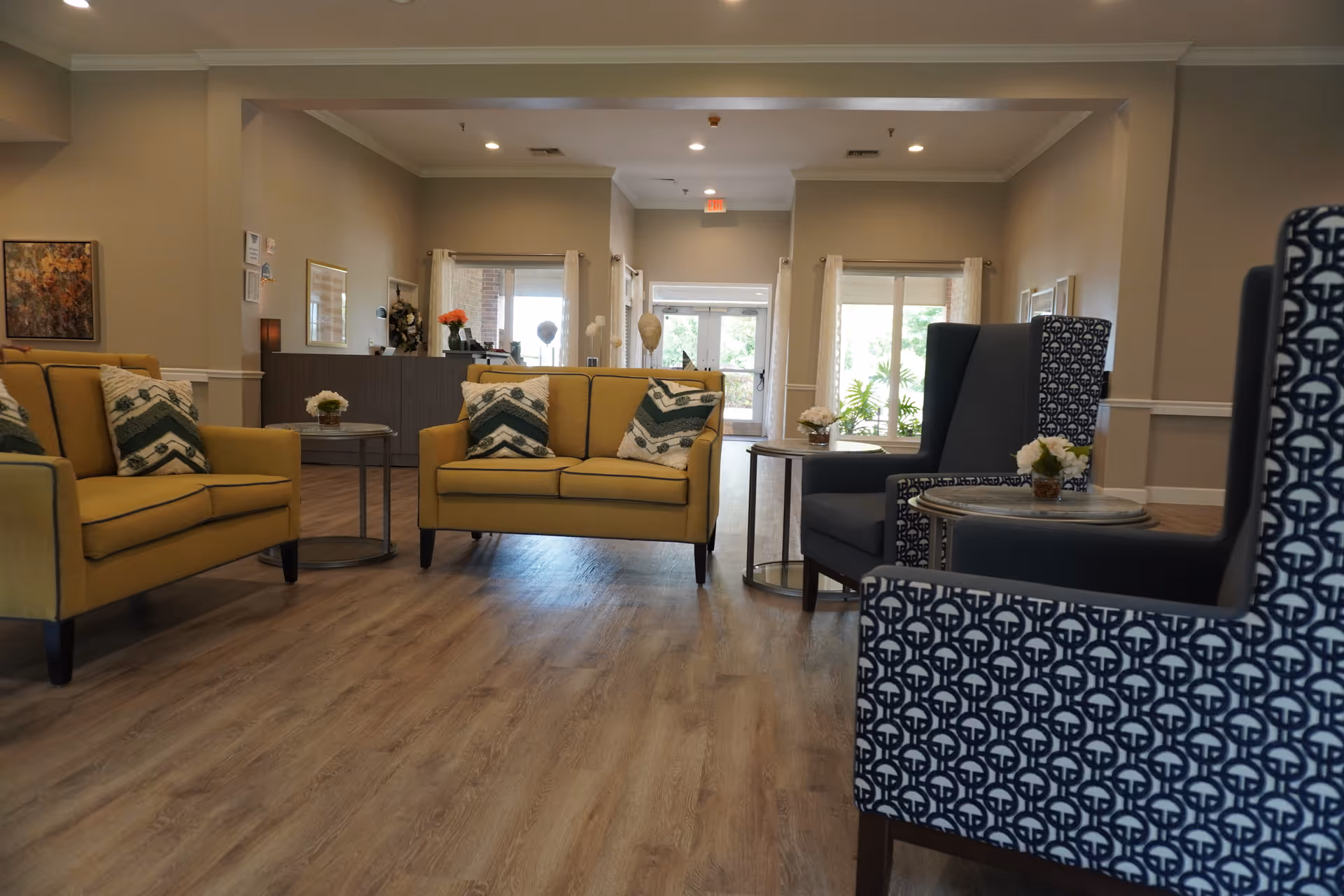 A spacious and well-lit living room area in a senior living facility featuring two yellow sofas with patterned cushions, two blue patterned armchairs, small round tables with flower arrangements, wooden flooring, and a reception desk in the background near large windows and glass doors.