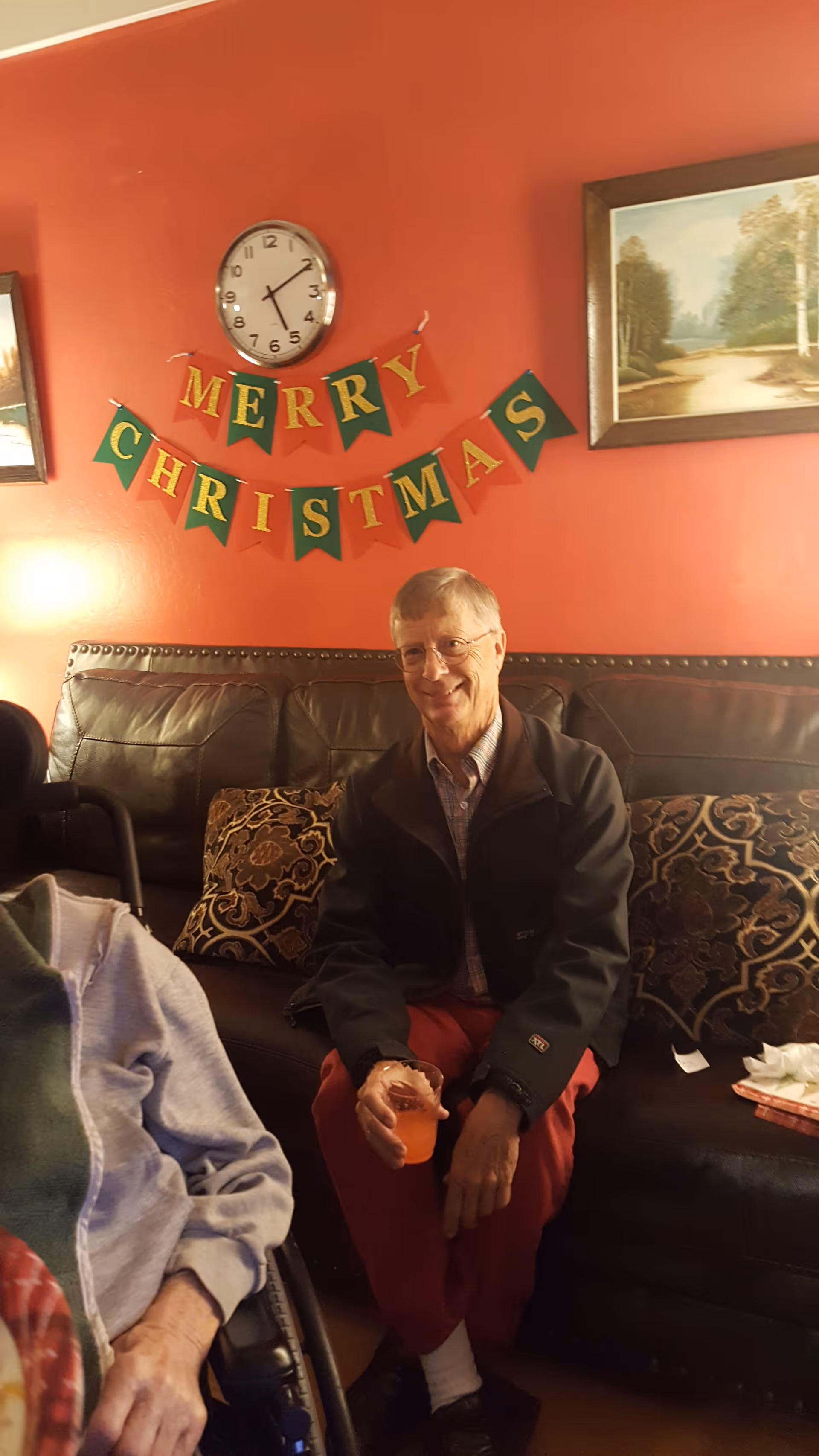 An elderly man sitting on a leather couch in a red-walled room decorated with a 'Merry Christmas' banner and framed paintings.