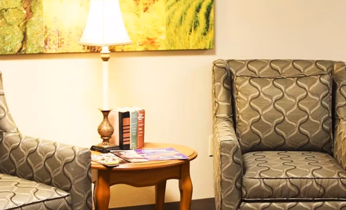Two patterned armchairs with a wooden side table between them. The table holds a lamp, several books, and some magazines. A colorful abstract painting is partially visible on the wall behind the lamp.