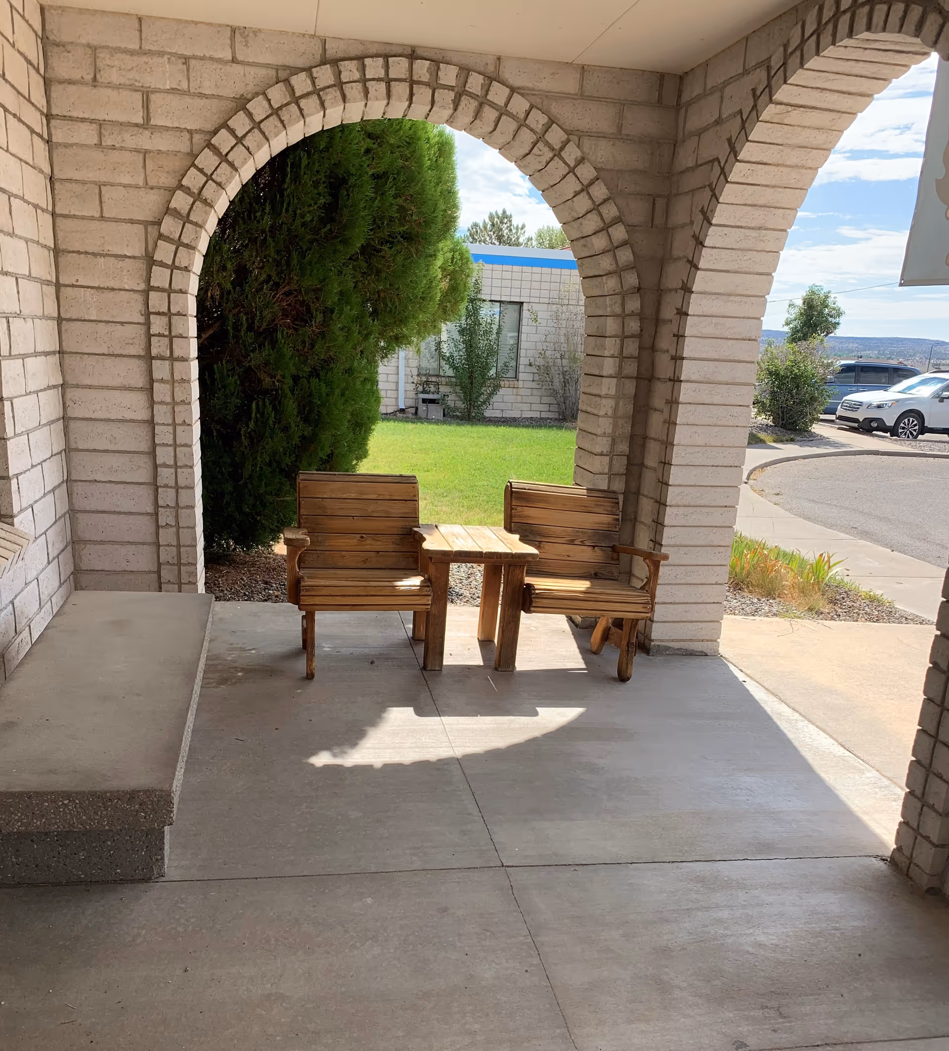 Two wooden chairs and a small table sit under brick archways on a covered porch overlooking a lawn and parked cars.