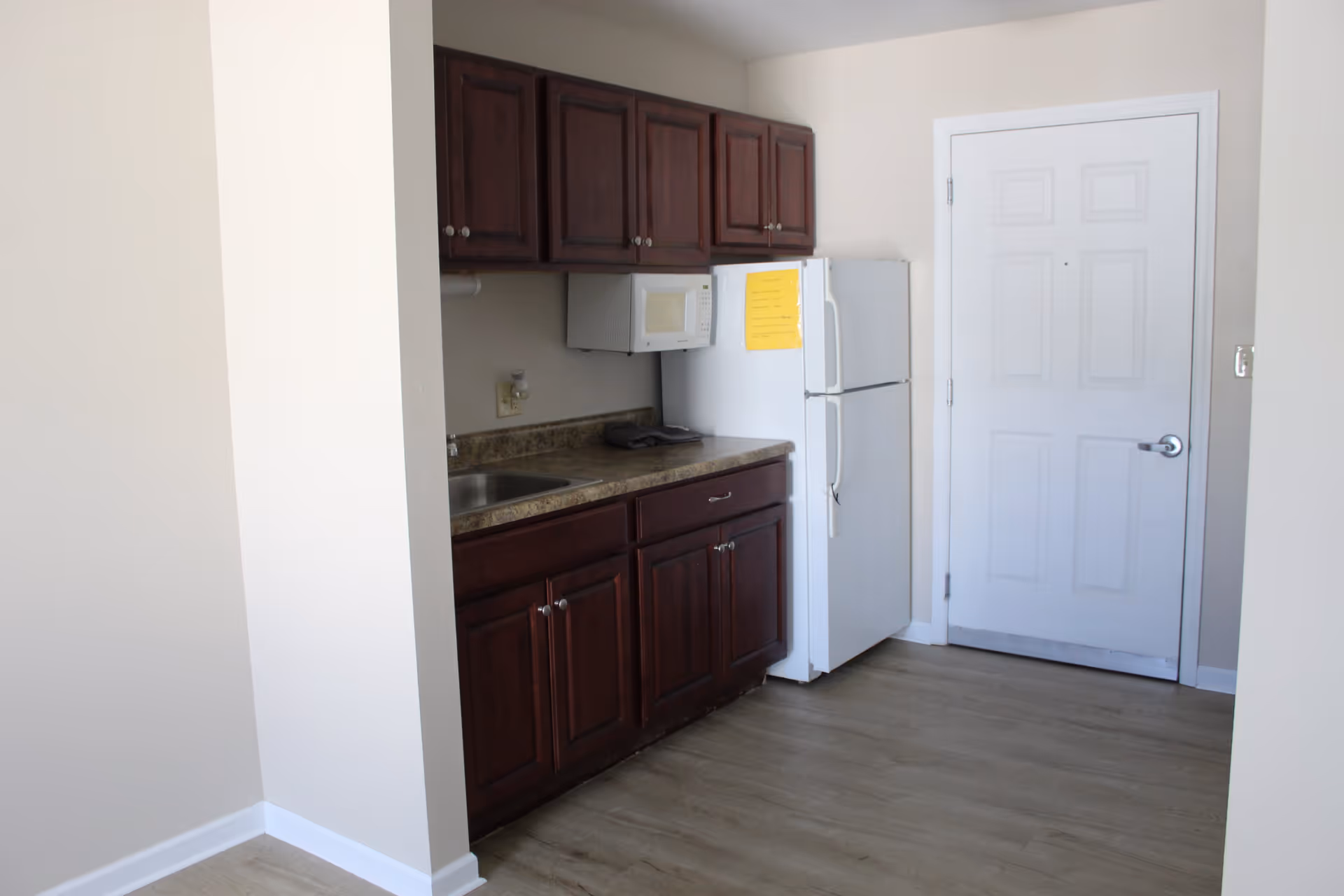 Small kitchen area with dark wooden cabinets, a countertop with a sink, a white microwave mounted above the counter, and a white refrigerator next to a closed white door. The floor is light wood and the walls are painted beige.