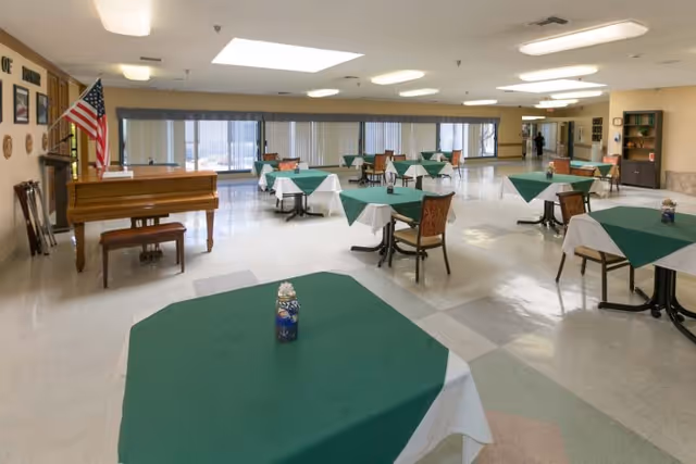 A spacious dining room with multiple tables covered with green and white tablecloths, each table having a small centerpiece. There are chairs around the tables, a piano with a bench, and an American flag in the corner. The room has large windows with vertical blinds and ceiling lights providing illumination.