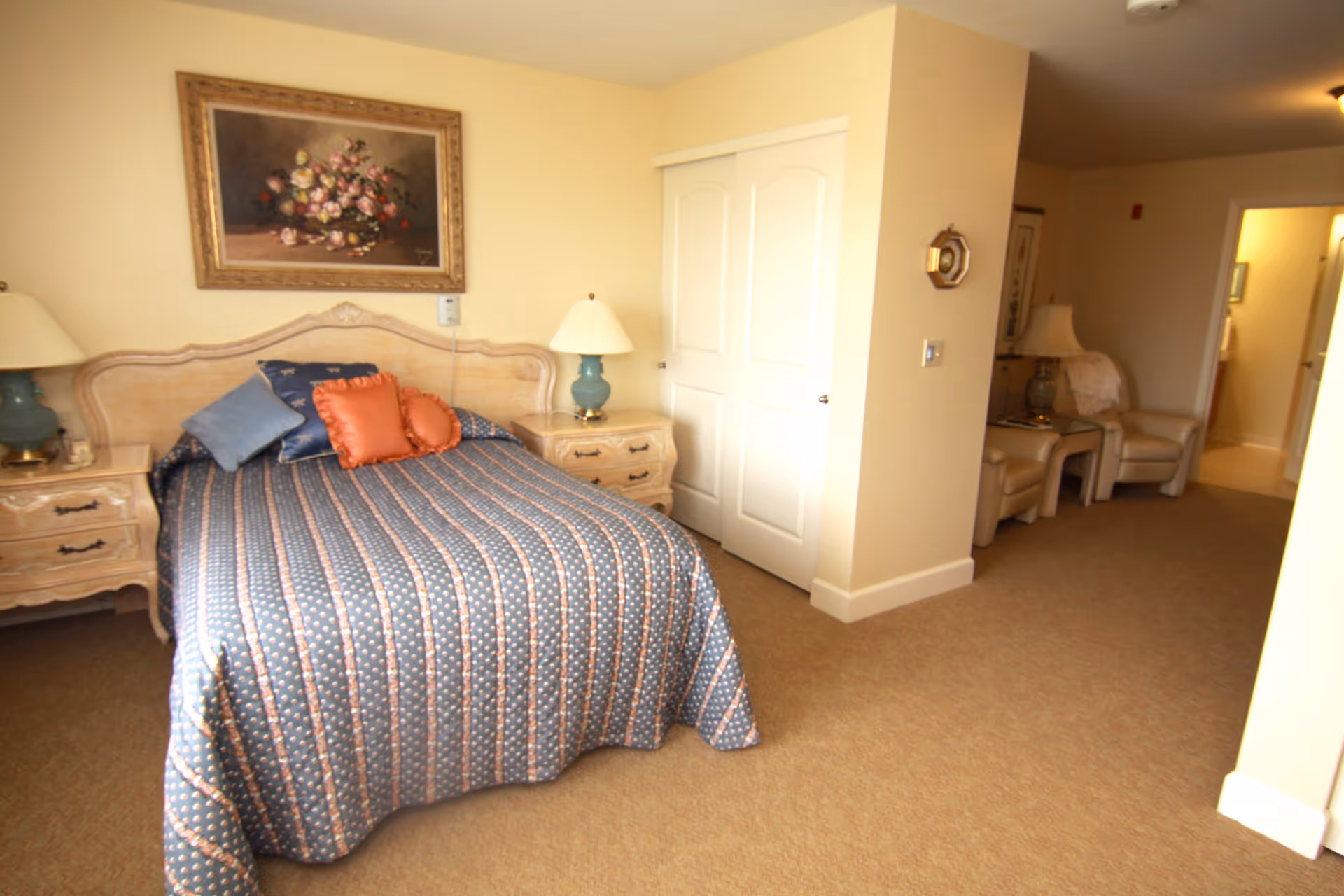 A bedroom with a bed covered in a blue patterned bedspread and several pillows. There are two matching nightstands with lamps on either side of the bed. A framed floral painting hangs above the headboard. In the background, there are two beige armchairs with a small table and lamp between them, and a hallway leading to another room.