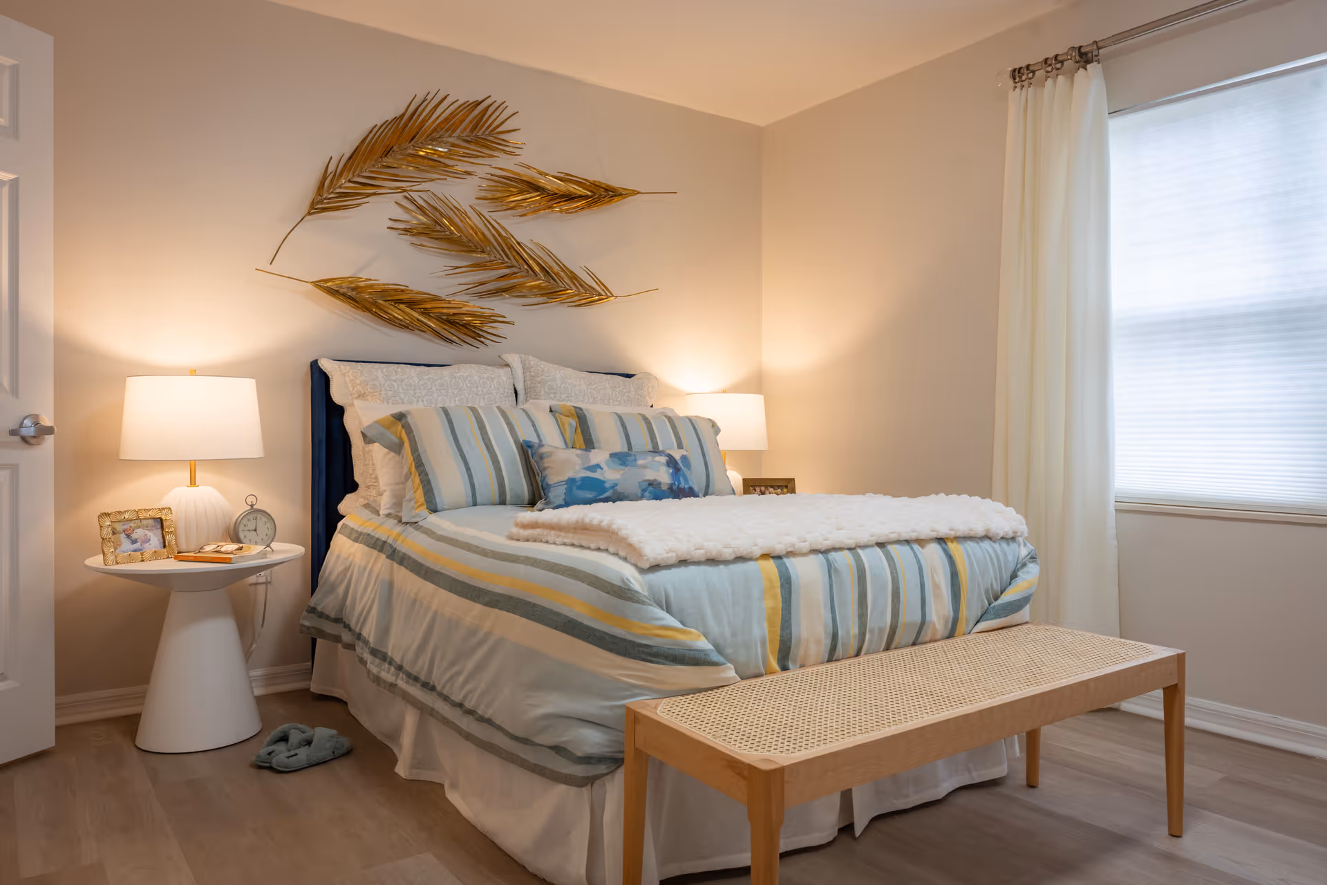 A cozy bedroom with a neatly made bed featuring striped bedding in shades of blue, yellow, and white. There are two bedside tables with lamps on each side of the bed. Above the bed, there is a decorative wall art piece made of golden palm leaves. A wooden bench is placed at the foot of the bed, and a window with white curtains allows natural light into the room.