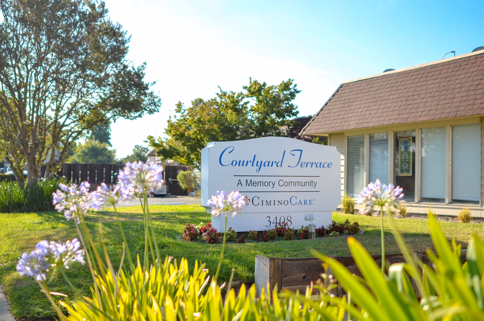 Outdoor view of Courtyard Terrace, a memory community facility, showing a white sign with blue and black text surrounded by green grass, flowering plants, and trees, with part of a building with windows and a brown roof in the background.