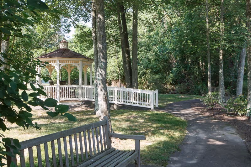 A peaceful outdoor garden area with a wooden bench in the foreground, a white gazebo with a shingled roof in the midground, surrounded by tall trees and greenery, and a paved walking path curving through the scene.