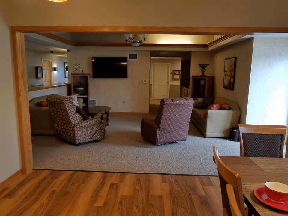 A cozy living room area in a senior living facility with two patterned armchairs, a beige sofa with cushions, a wall-mounted flat-screen TV, wooden cabinets, and a framed picture on the wall. The room has carpeted flooring and is viewed from a dining area with wooden flooring and a table with chairs.