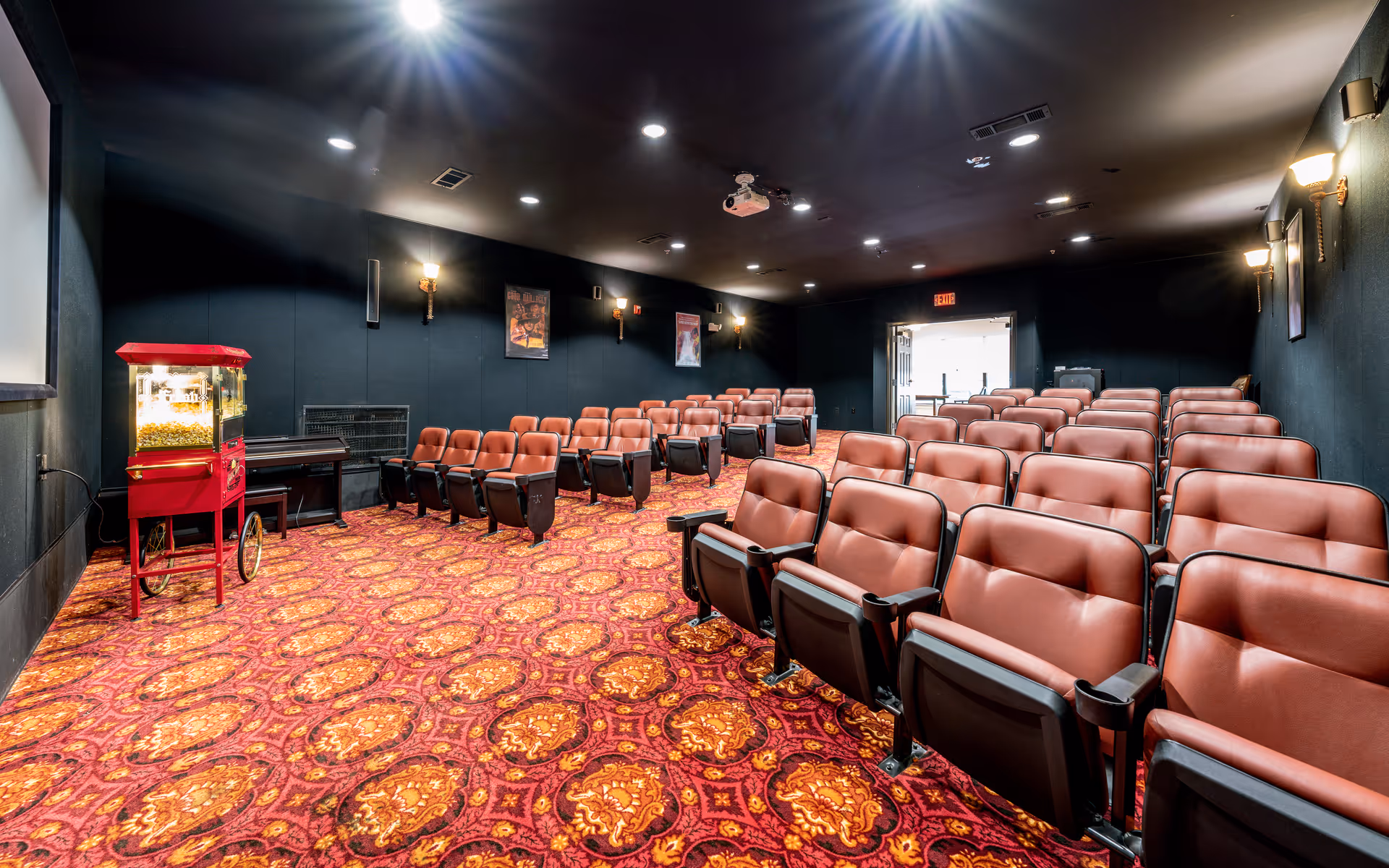 Interior view of a small movie theater room with rows of red cushioned seats, a red popcorn machine on the left side near the screen, patterned red and orange carpet, dark walls, and ceiling lights.
