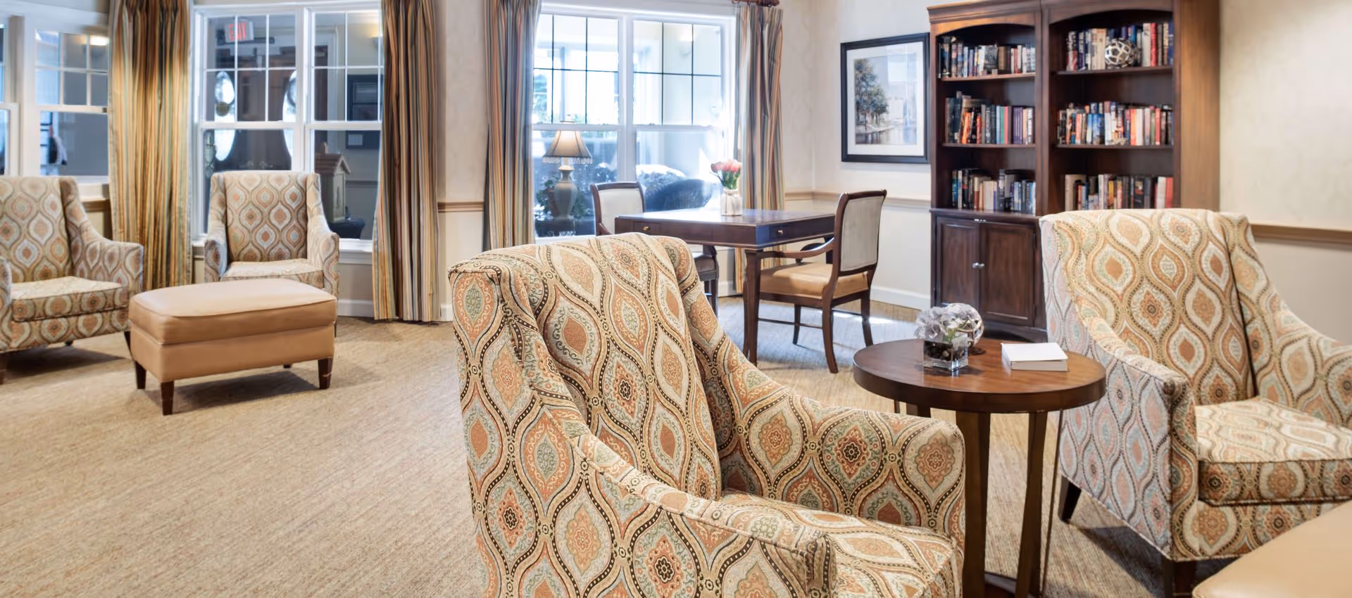 A bright senior living common room with patterned armchairs, a round side table, bookshelf, and windows with curtains.