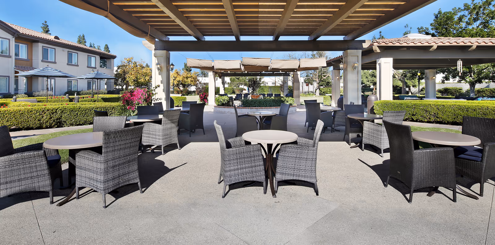 Outdoor patio area with multiple round tables and wicker chairs under a pergola. Surrounding the patio are well-maintained hedges, flowering plants, and trees. In the background, there are buildings with tiled roofs and windows, and a clear blue sky above.