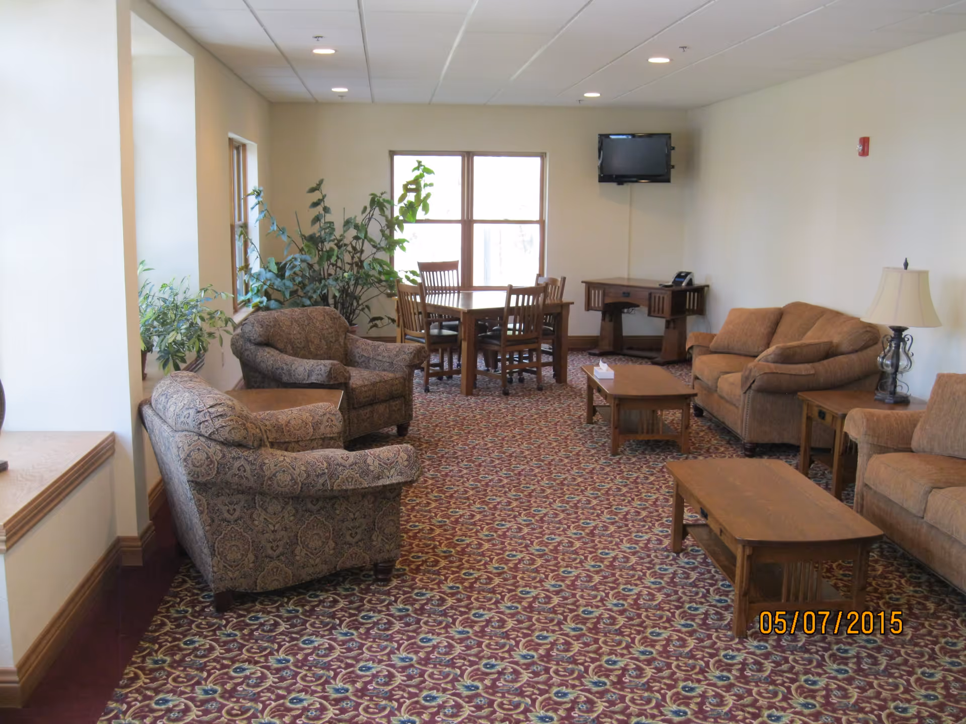 A cozy living room area with patterned carpet, two upholstered armchairs, a brown sofa, a wooden coffee table, a side table with a lamp, and a wooden dining table with four chairs near a window. There are also some potted plants and a wall-mounted TV in the corner.
