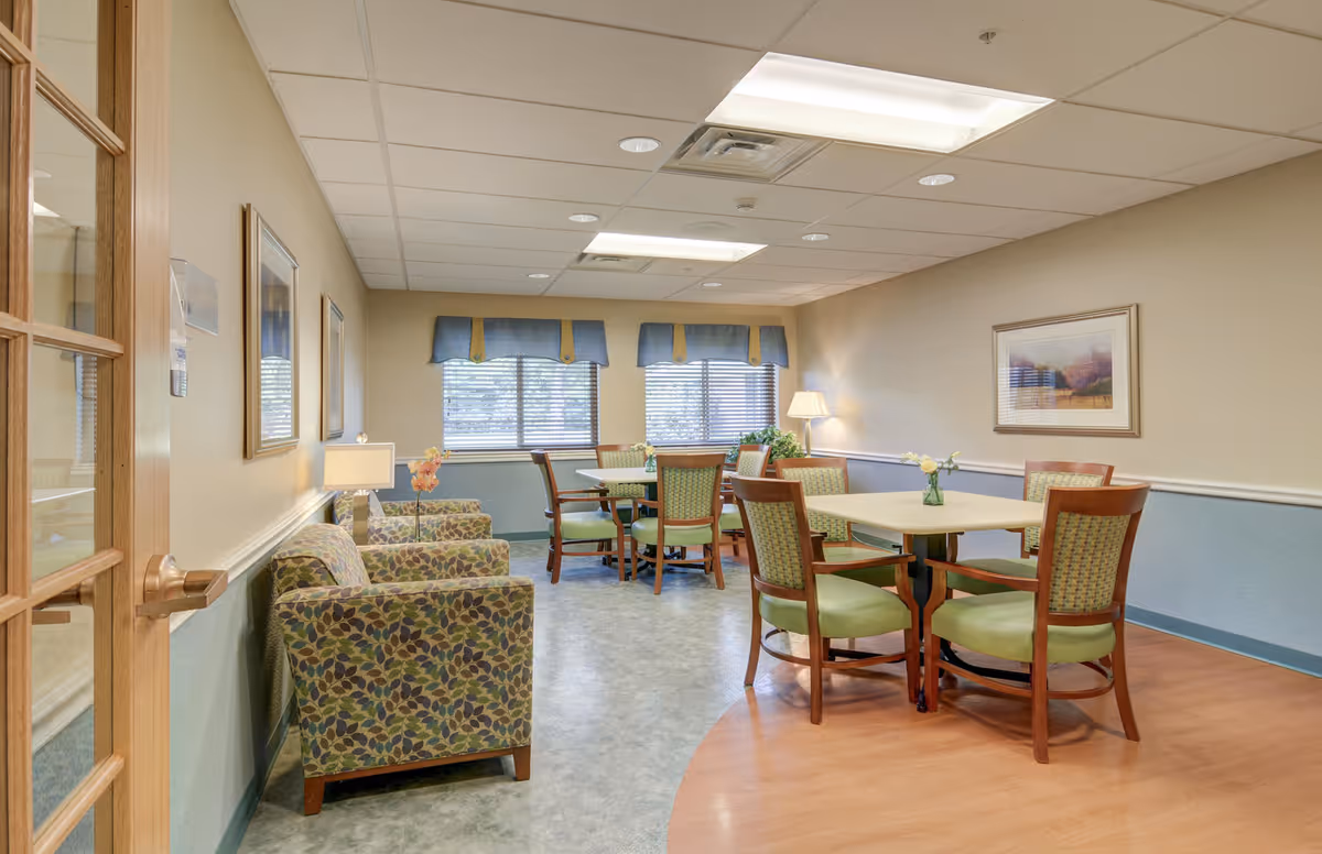Bright senior living common room with round dining tables and green-cushioned chairs, patterned armchairs, lamps, and windows with blue valances.