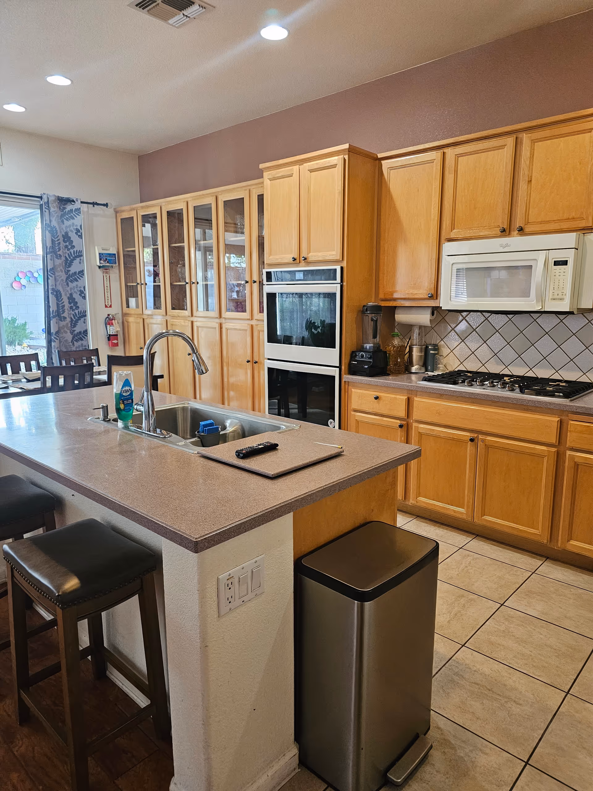 A modern kitchen with wooden cabinets, a built-in oven, a microwave above a gas stove, and a kitchen island with a sink and two bar stools. There is a stainless steel trash can next to the island, and a dining area with chairs and a sliding glass door leading outside is visible in the background.