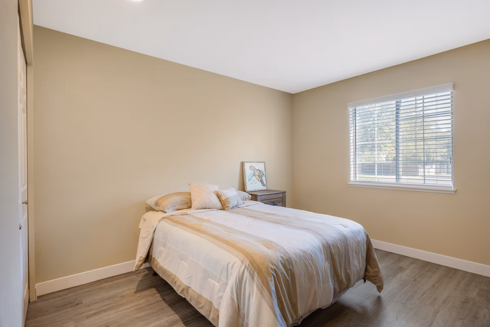 Simple bedroom with a made bed, nightstand, window with blinds, and beige walls.