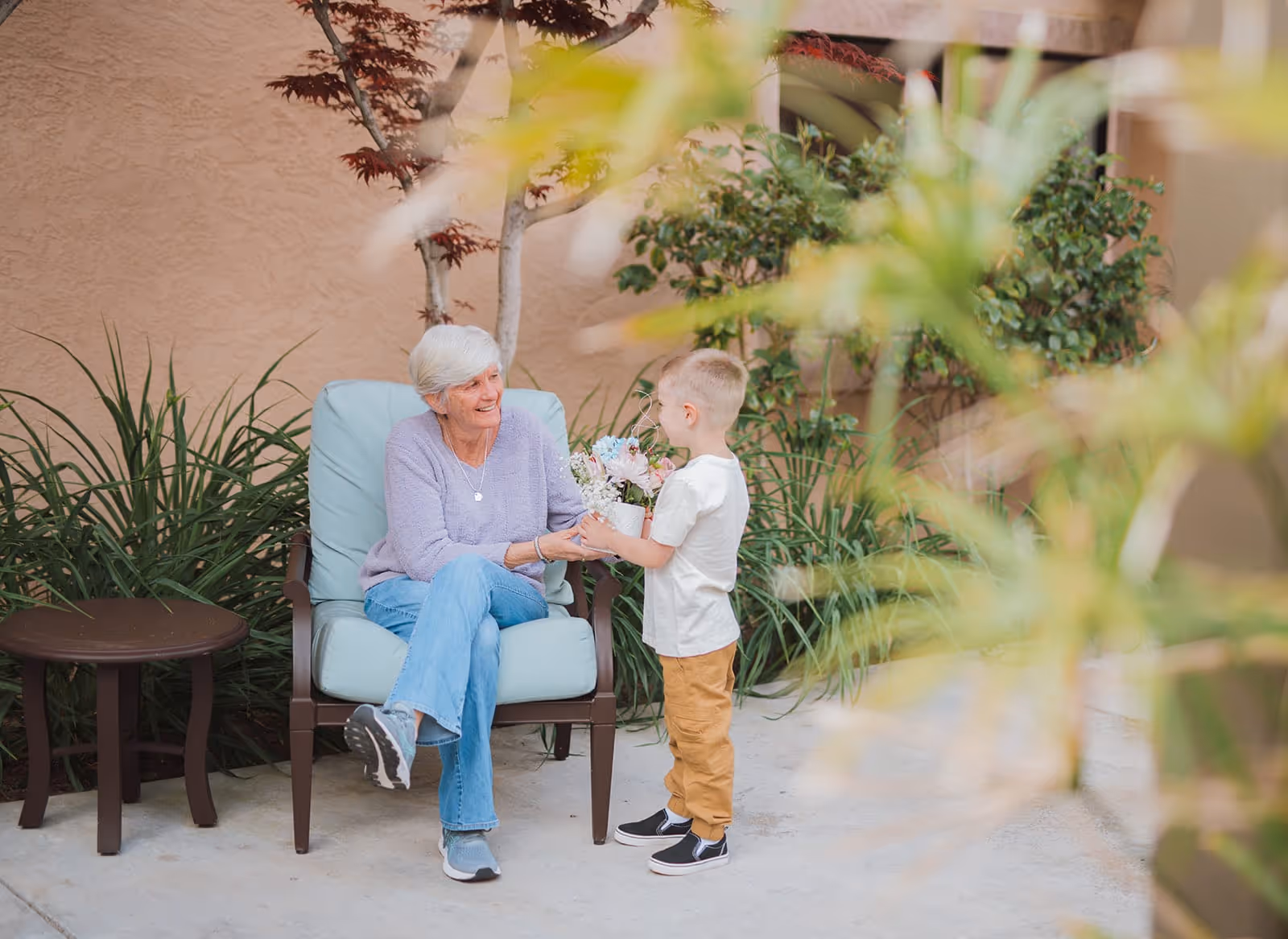 An elderly woman with gray hair sitting on a cushioned outdoor chair smiling and receiving a flower pot from a young boy standing in front of her. They are in a garden area with plants and a small table nearby.