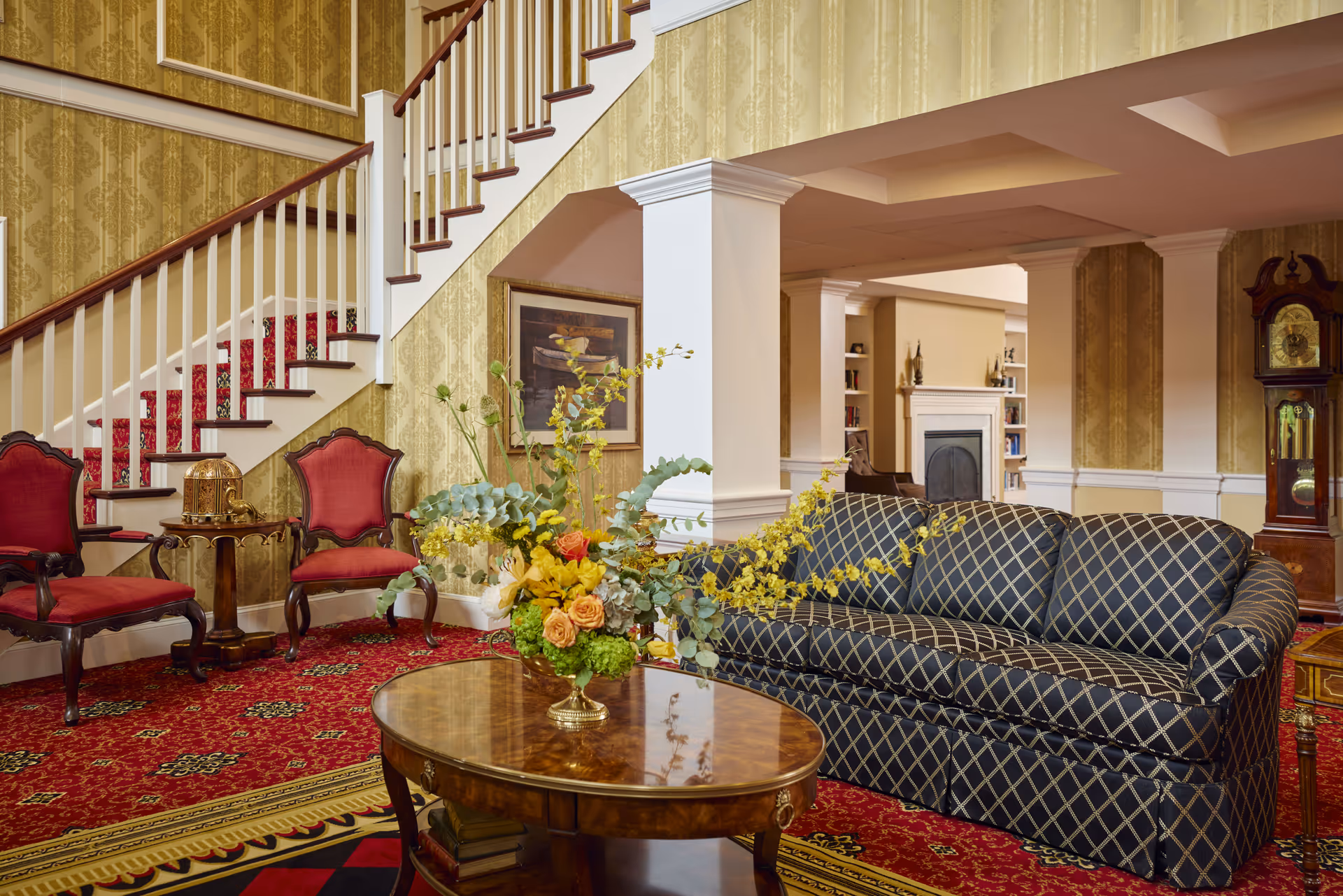 A cozy and elegant living room area with a patterned dark blue sofa, a wooden coffee table with a floral arrangement, two red upholstered chairs, and a staircase with a red carpet runner. The room features yellow patterned wallpaper, a grandfather clock, and a fireplace in the background.