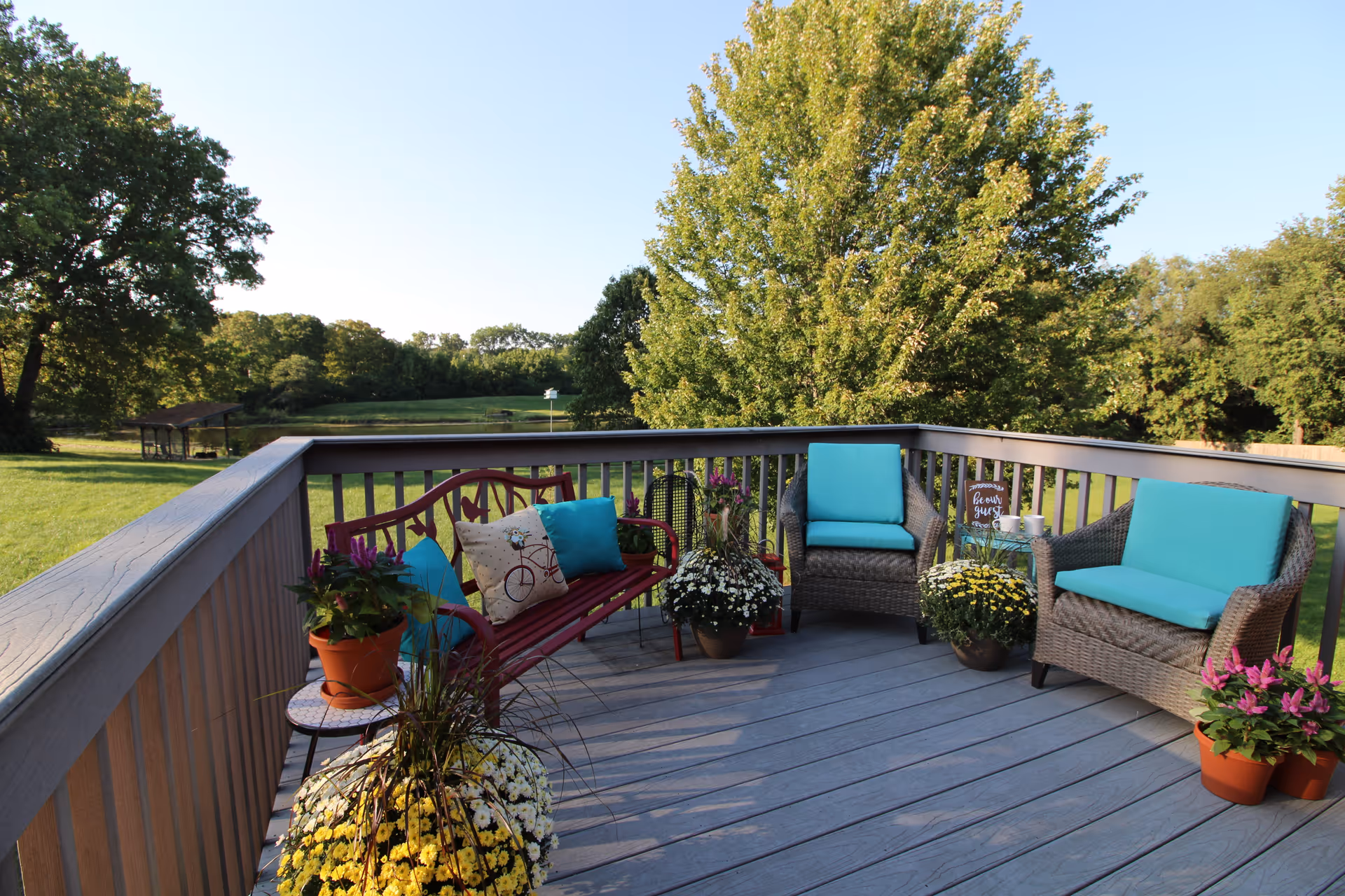 Outdoor deck area with a red bench and two wicker chairs with turquoise cushions, surrounded by potted flowers and plants, overlooking a grassy yard with trees and a clear blue sky.