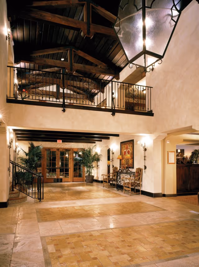 Spacious interior hallway with a high wooden beam ceiling, wrought iron railing on the upper level, large hanging lantern light fixture, tiled floor, potted plants, decorative chairs, and a console table with a lamp and framed artwork on the wall.