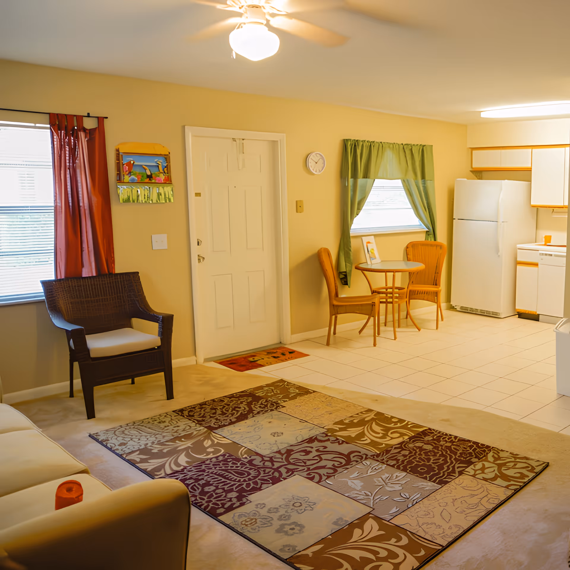 Open-plan living area with a patterned area rug, chairs and a small round dining table by a window, and a kitchenette with a refrigerator.