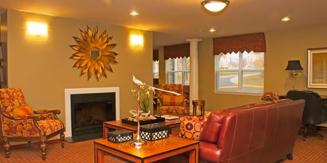 A cozy living room with a red leather sofa, two patterned armchairs, a wooden coffee table with decorative items, a fireplace with a sunburst mirror above it, warm lighting, and windows with valance curtains showing an outdoor view.