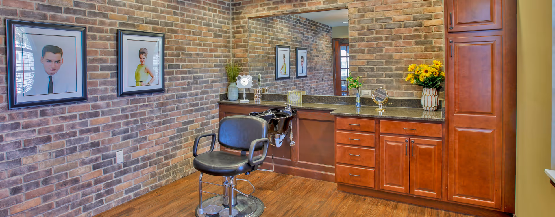 Interior of a hair salon area with a black salon chair in front of a black sink. The walls are covered with brown brick wallpaper and decorated with framed vintage portraits. There is a large mirror above a dark granite countertop with wooden cabinets below. The countertop has decorative items including a vase with yellow flowers, a small clock, and a small mirror.