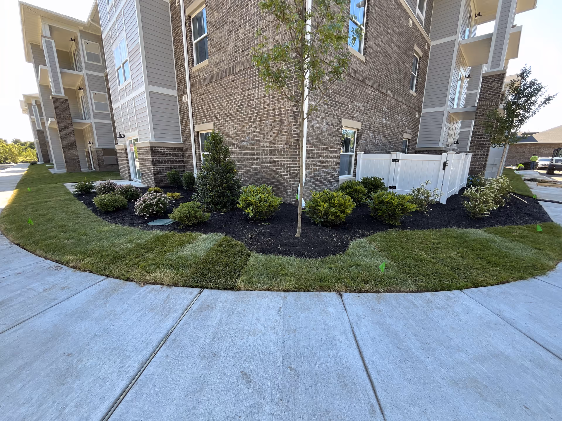 Landscaped corner of a multi-story brick and siding retirement building with a sidewalk and a small white fenced patio.