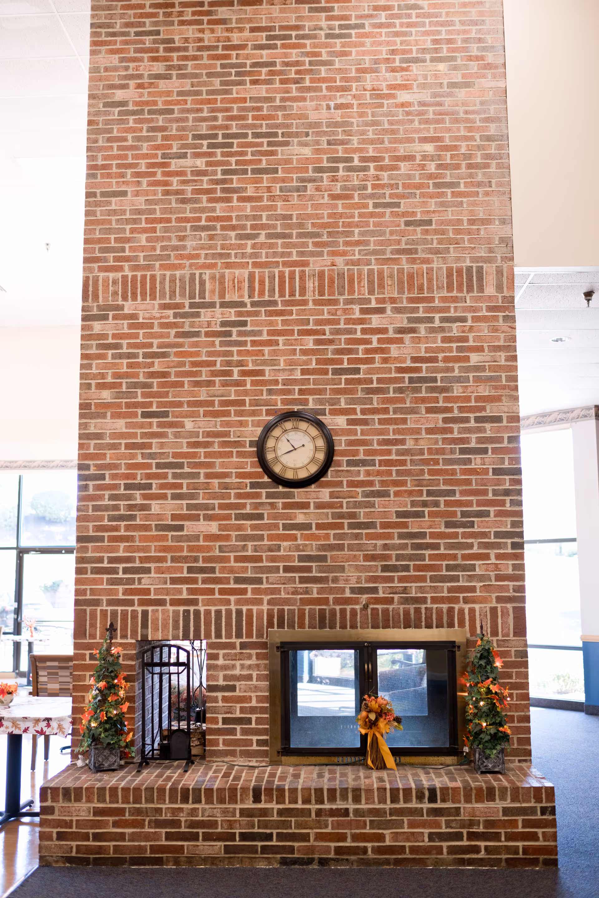 A tall brick fireplace with a black-framed clock mounted above it. The fireplace has glass doors and is decorated with small artificial trees with orange lights and a fall-themed floral arrangement. The surrounding area includes large windows letting in natural light and a table with a floral tablecloth partially visible on the left.