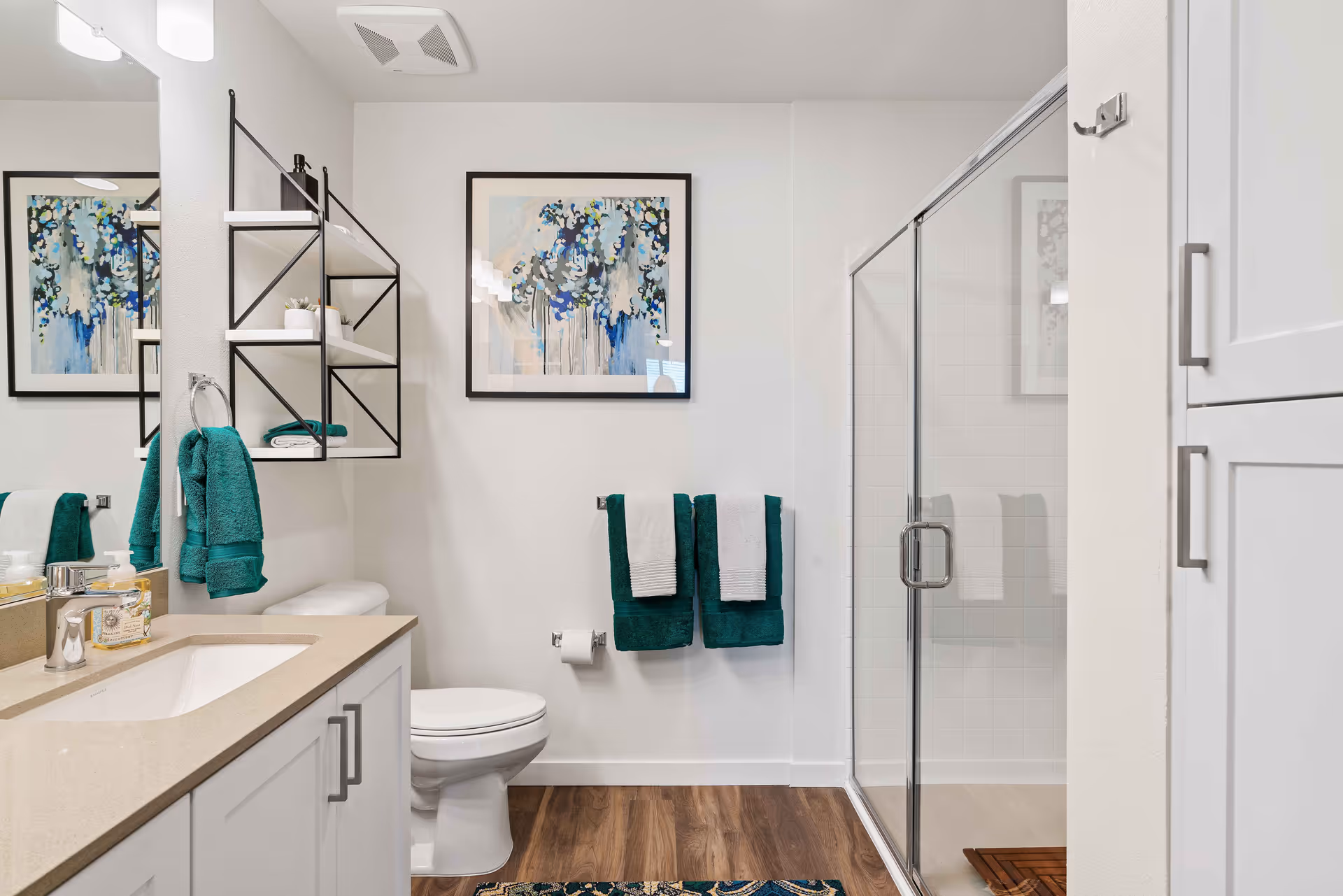 A modern bathroom with a white vanity and beige countertop, a toilet, a glass-enclosed shower, and white walls. There are teal and white towels hanging on racks, a black metal wall shelf with decorative items, and a framed floral artwork on the wall. The floor is wood-style, and there is a patterned rug in front of the vanity.