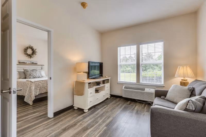 A cozy living room with a gray couch adorned with pillows, a white TV stand holding a flat-screen television and a lamp, and two windows with white blinds letting in natural light. The room has wood flooring and cream-colored walls. An open door reveals a bedroom with a bed decorated with patterned bedding and a wreath hanging above the headboard.