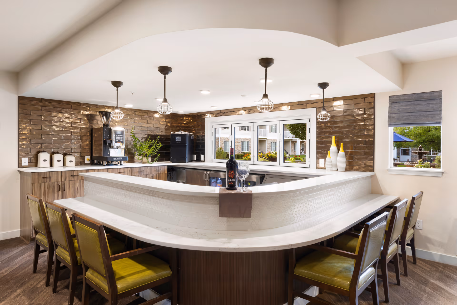 A modern kitchen area in a senior living facility with a white curved countertop bar surrounded by six chairs with green cushions. The kitchen features wooden cabinets, a coffee machine, a black appliance, decorative vases, and three hanging pendant lights. There are windows showing an outdoor garden area.