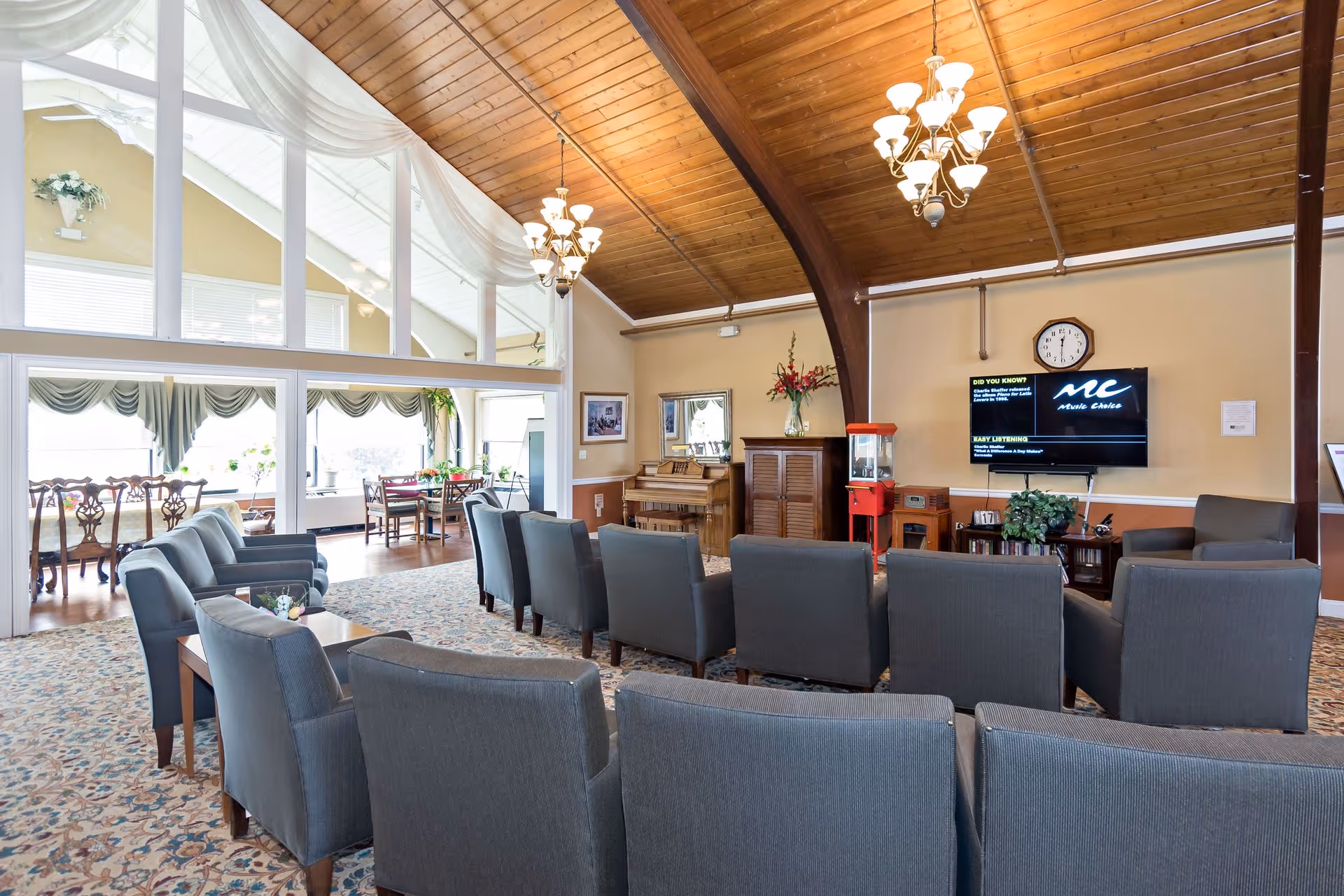A spacious living room area with multiple gray armchairs arranged in rows facing a wall-mounted TV. The room features a high wooden vaulted ceiling with chandeliers, a patterned carpet, and a piano against the far wall. Adjacent to the living room is a dining area with wooden chairs and tables near large windows with draped curtains, allowing natural light to fill the space.
