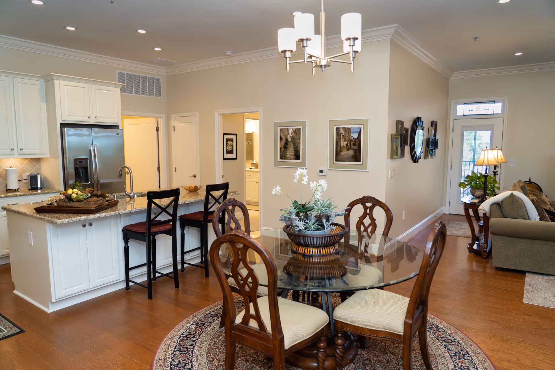 Interior view of a senior living facility showing a kitchen with white cabinets, stainless steel refrigerator, and a granite countertop island with two bar stools. In the foreground, there is a round glass dining table with four wooden chairs on a patterned rug. The background includes a living area with a sofa, side table with a lamp, and a door leading outside. The walls are decorated with framed pictures and a mirror.