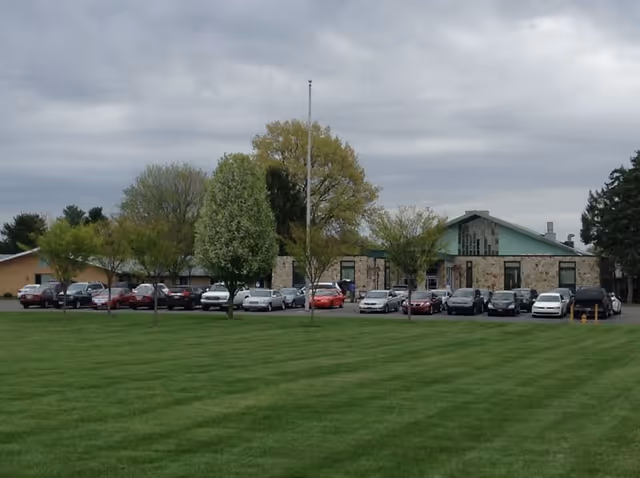 A single-story building with a stone facade and green roof surrounded by a parking lot filled with cars. Several trees are planted in front of the building, and a large grassy lawn is in the foreground under a cloudy sky.