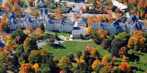 Aerial view of Cordia at Grand Traverse Commons, a large multi-wing building surrounded by trees with autumn foliage and green lawns, with parking areas visible in the background.