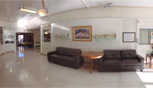 A spacious interior common area with two dark brown leather sofas separated by a round wooden table. The walls are light-colored with decorative molding and a framed painting above the sofas. There are wall-mounted candle holders with green garlands on either side of the painting. The floor is light-colored tile, and the ceiling has fluorescent and pendant lighting. In the background, there is a hallway leading to another room with a piano visible.