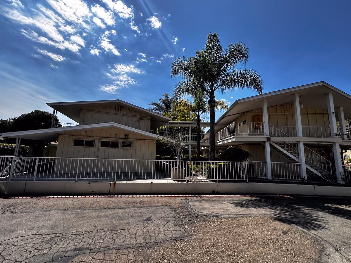 Exterior front view of a two-story senior living building with balconies, palm trees, and a paved driveway under a blue sky.
