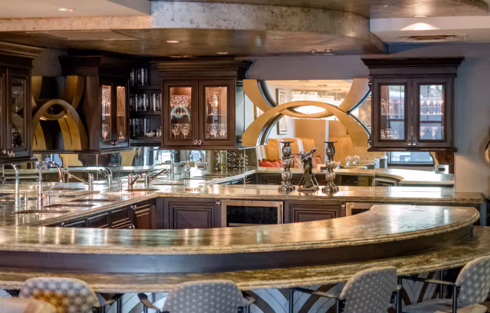 A luxurious kitchen area with a curved marble countertop and dark wooden cabinets with glass doors displaying glassware. The kitchen features multiple sinks and faucets, with a mirrored backsplash reflecting the room. Decorative candle holders and a small statue are placed on the countertop. In the background, a living area with beige seating and a large circular mirror with a unique design is visible.