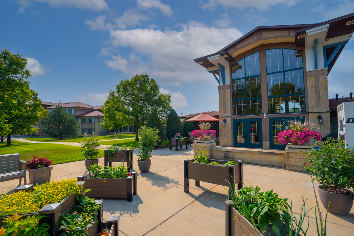 Outdoor courtyard area at Meadows on Fairview featuring raised garden beds with various plants and flowers, benches, large trees, and a building with large windows and stone accents under a partly cloudy blue sky.