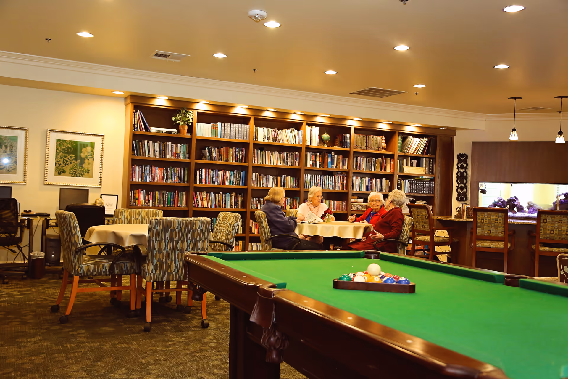A cozy common area in a retirement community featuring a pool table in the foreground, a round table with four elderly women seated and conversing, bookshelves filled with books along the back wall, and additional seating and computer stations on the left side.