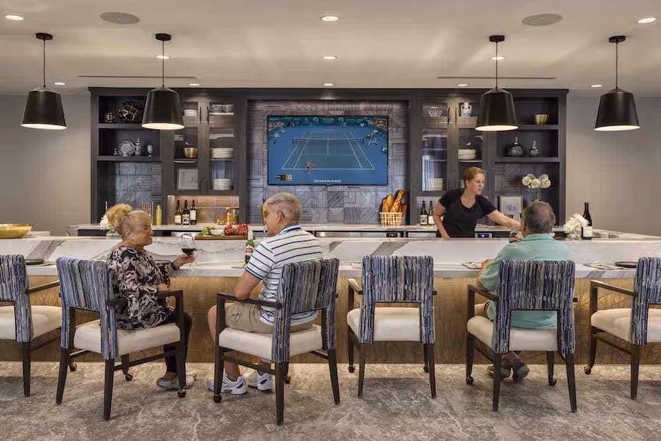 A modern bar area inside Knollwood Place Apartments with three senior individuals seated on cushioned chairs at a marble countertop. A bartender is serving drinks behind the counter. The background features built-in shelves with decorative items and a large flat-screen TV displaying a tennis match. The space is well-lit with pendant lights hanging from the ceiling.
