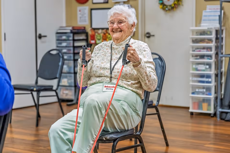 An elderly woman with white hair and glasses is sitting on a chair indoors, smiling while exercising with red resistance bands. She is wearing a floral long-sleeve shirt and light green pants, with a name tag that reads 'Ambassador Shirley'. The room has wooden floors, chairs, and storage drawers in the background.