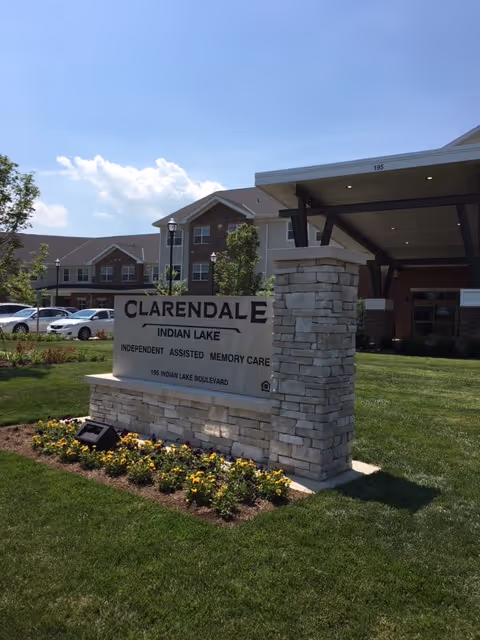 Outdoor view of the entrance area of Clarendale at Indian Lake senior living facility, showing a stone sign with the facility name and services offered, surrounded by green grass and flowers, with the building and parked cars in the background under a blue sky.