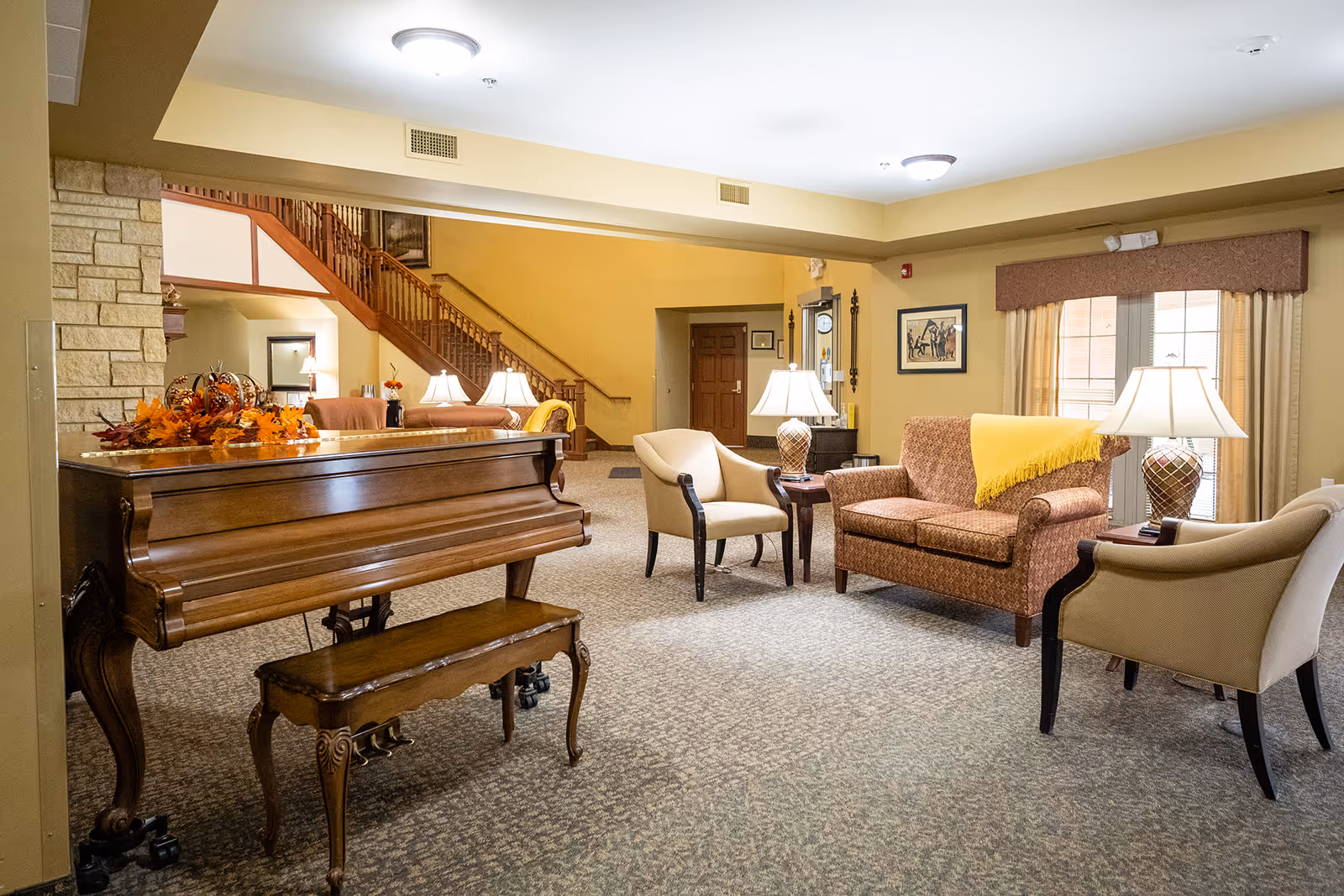 Bright communal living room with a wooden piano, armchairs, a loveseat with a yellow throw, table lamps, and a staircase in the background.