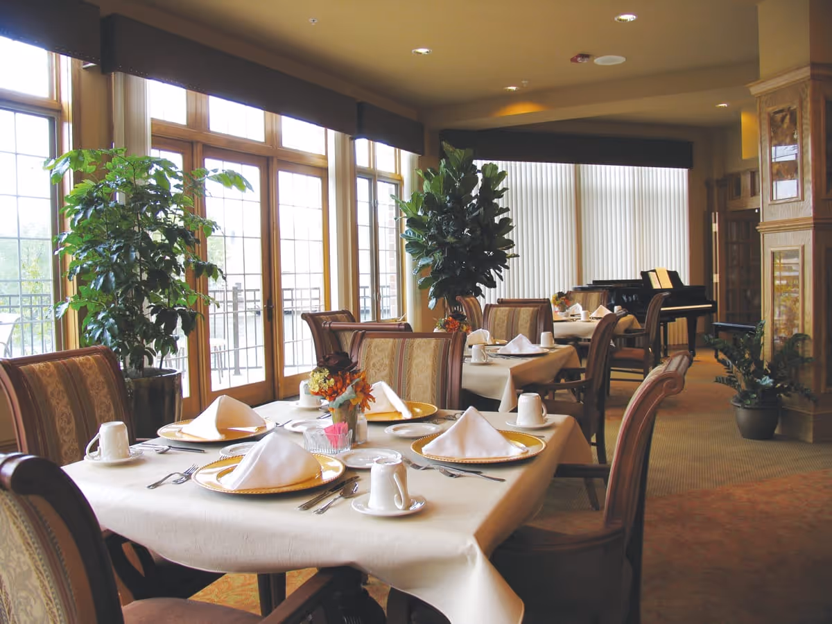 A dining room with tables set for a meal, featuring white tablecloths, folded napkins on plates, cups, and silverware. The room has large windows with wooden frames letting in natural light, potted plants, upholstered chairs, and a piano in the background.
