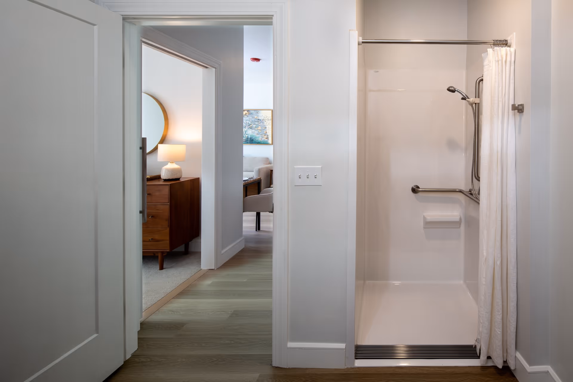 View of a walk-in shower with a white curtain and grab bars inside. To the left, there is an open doorway leading to a room with a wooden dresser, a round mirror, and a lit table lamp. Further in the background, a living area with a sofa and a chair is partially visible.