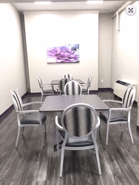 A small dining area with two square tables and eight silver chairs with black cushioned seats. The room has light-colored walls, dark wood flooring, a purple flower painting on the far wall, and a white heating or air conditioning unit on the right side.