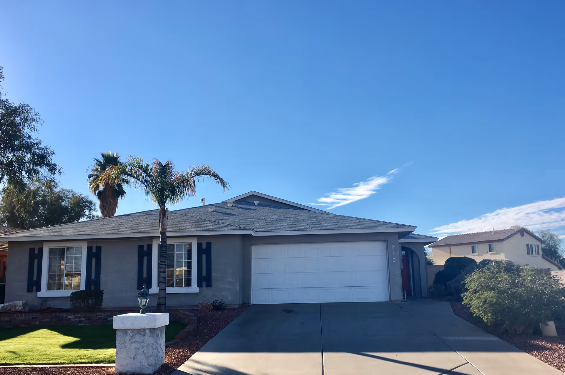Single-story suburban home front with an attached two-car garage, driveway, palm trees, and a clear blue sky.