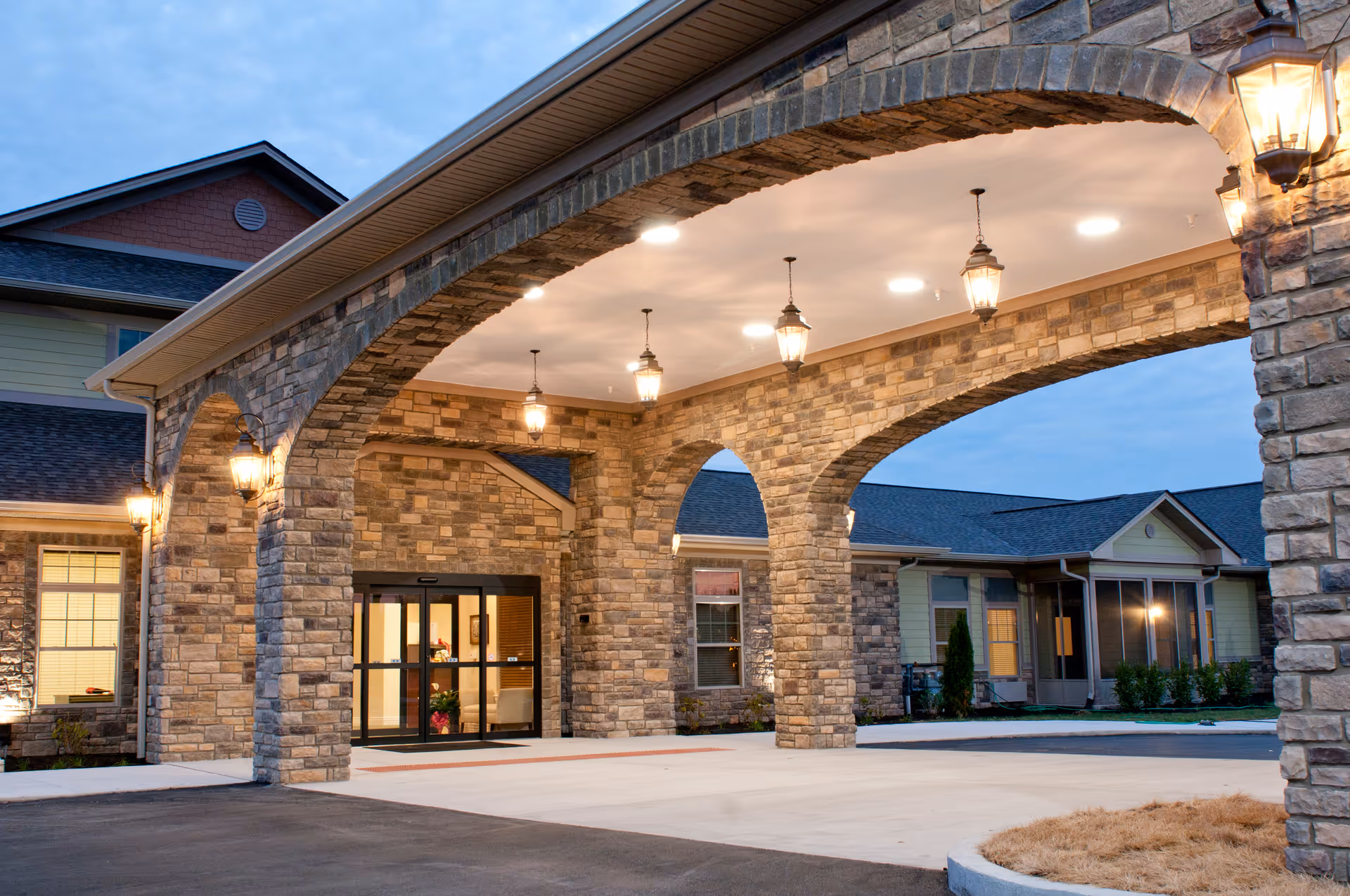 Entrance of a senior living facility with stone archways and hanging lantern-style lights, showing a covered driveway and glass doors leading inside the building during early evening.